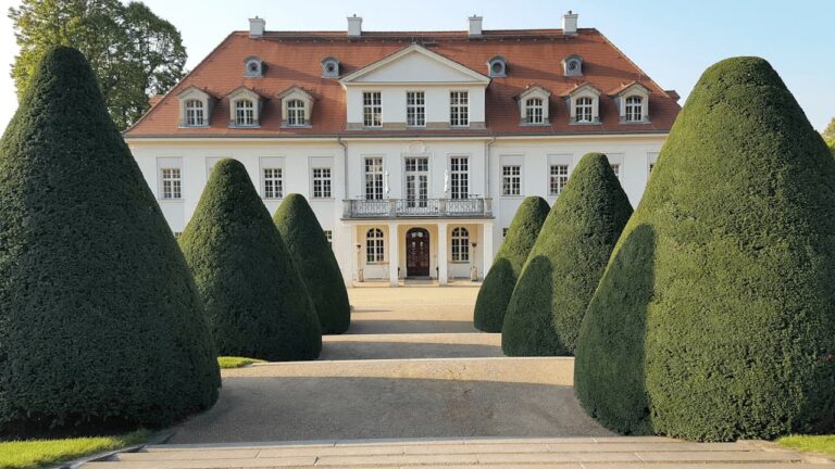Elegant baroque mansion with red-tiled roof behind formal garden featuring large cone-shaped topiary hedges requiring regular maintenance