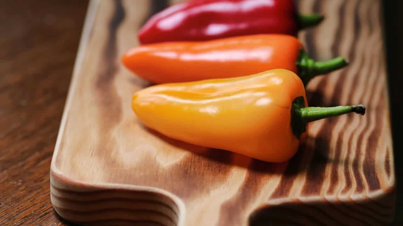 Three colorful mini sweet peppers in red, orange, and yellow arranged on a wooden cutting board with natural wood grain