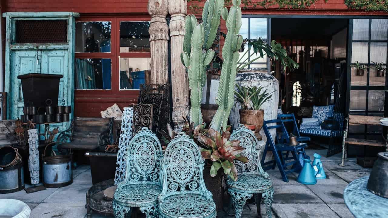 Cluttered antique patio with ornate metal chairs, large cactus, wooden columns, pottery, and various vintage items displayed