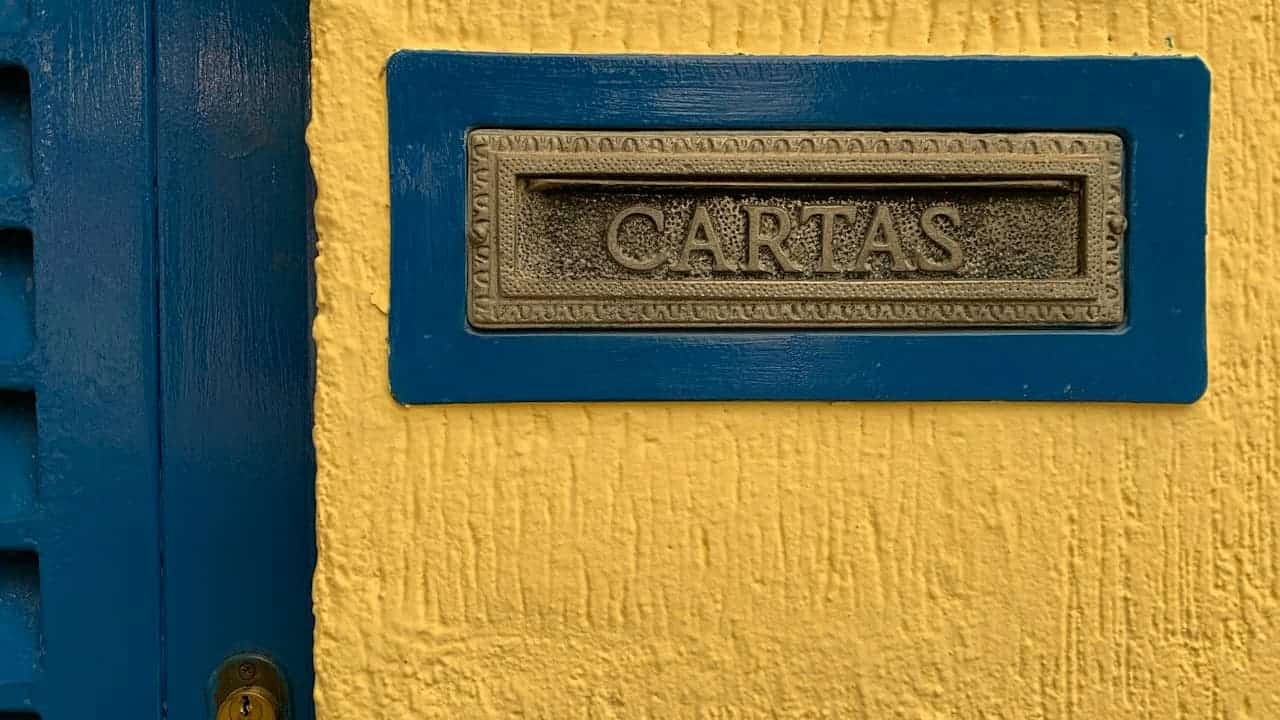Ornate brass mail slot labeled "CARTAS" mounted on blue wooden frame against textured yellow stucco wall