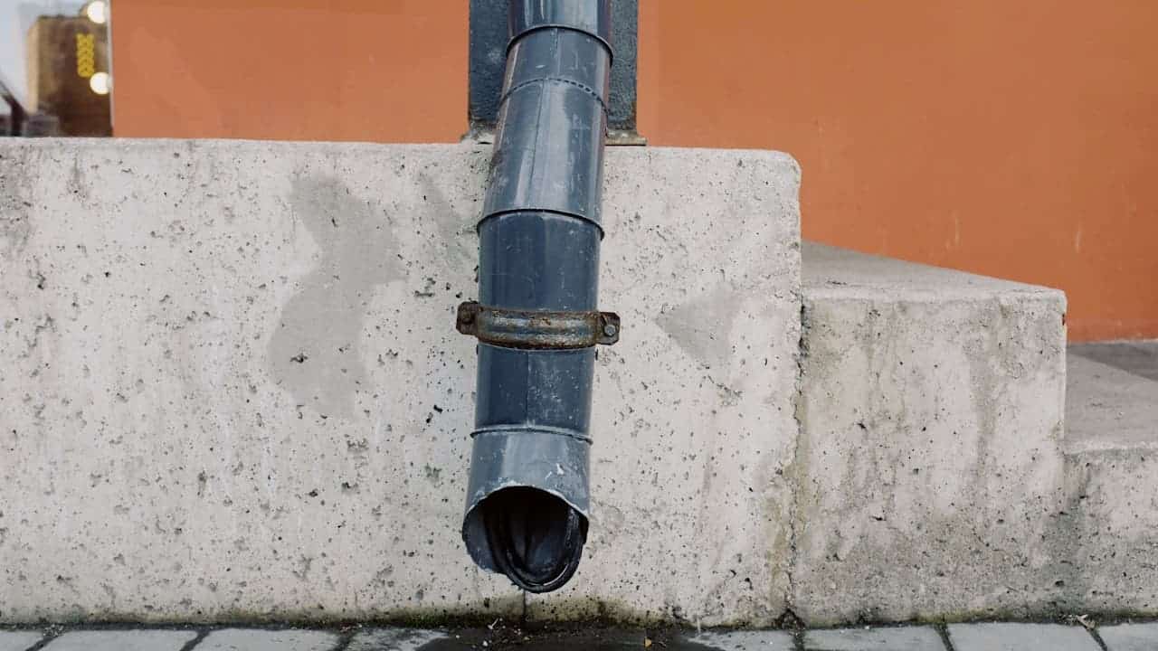 Gray metal downspout extending through concrete retaining wall with open end draining directly onto paved surface below