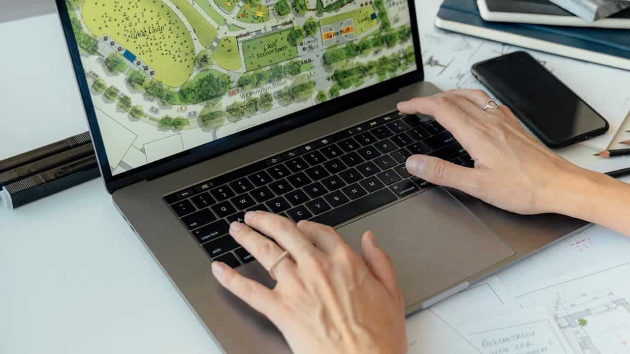 Person typing on laptop displaying aerial map view, hands positioned on keyboard, smartphone and papers visible on white desk
