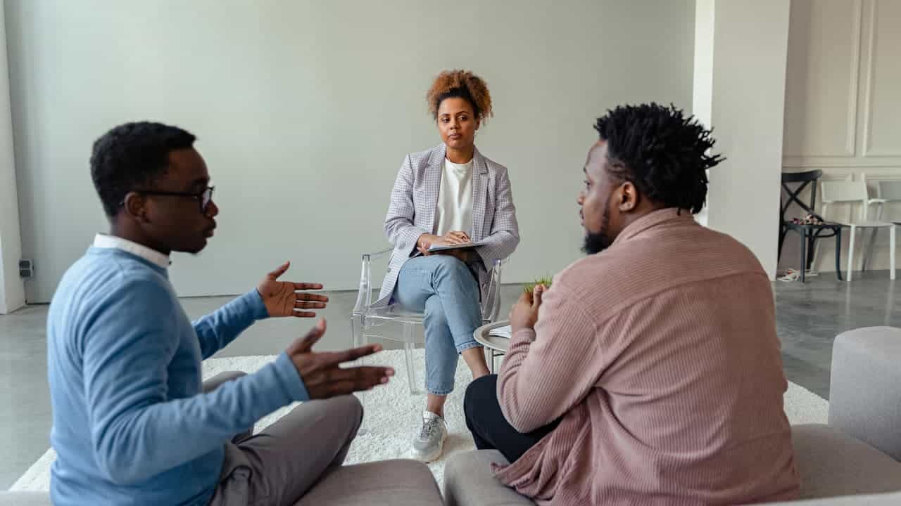 Two men and one woman seated indoors, actively mediating a discussion, facing each other in a calm setting