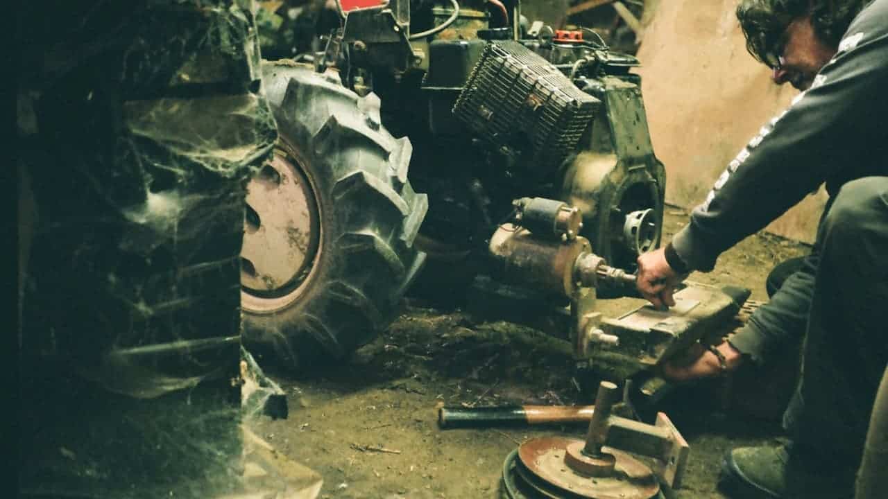 Mechanic working on disassembled tractor engine with large tires and mechanical components in workshop setting
