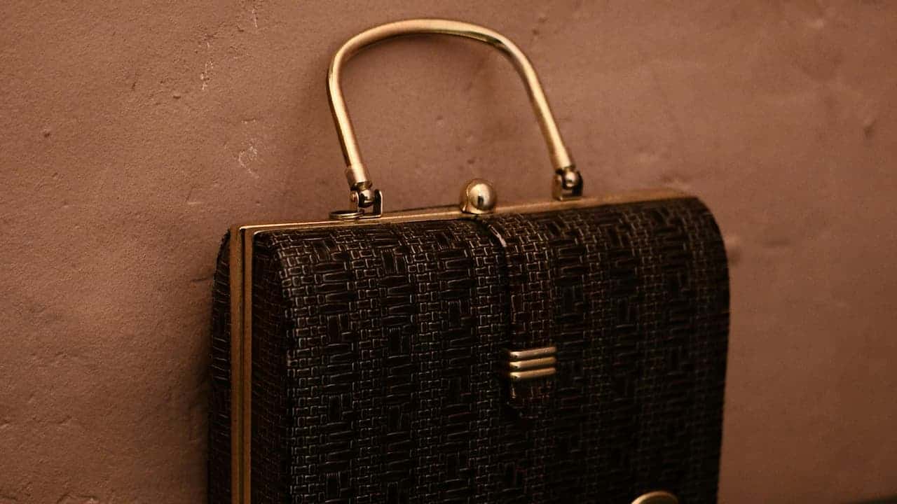  Dark textured handbag with gold metal frame and handle, hanging against a brown textured wall