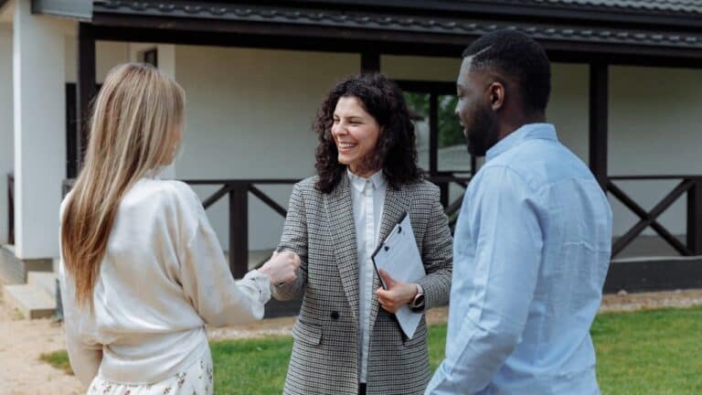 : A real estate agent in a checkered blazer shaking hands with clients outside a modern house