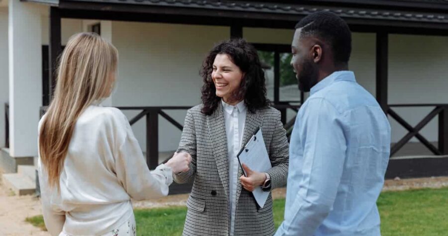 : A real estate agent in a checkered blazer shaking hands with clients outside a modern house