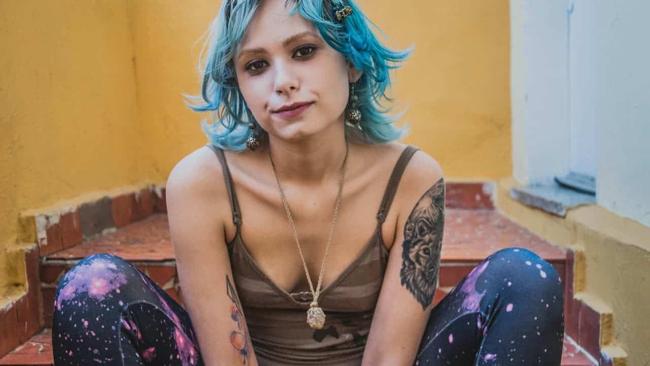 Young woman with blue hair sitting on tiled stairs, wearing galaxy-print leggings featuring a vivid cosmic pattern of pink