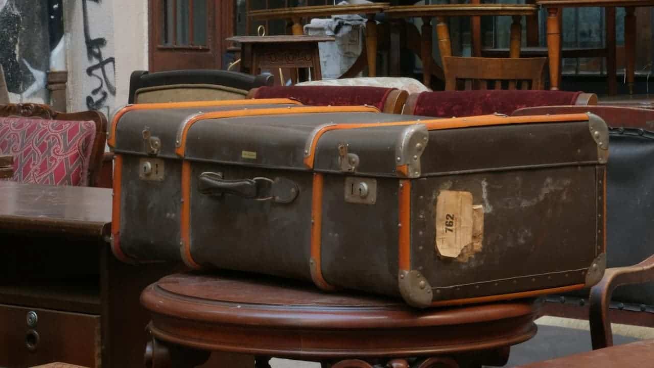 Worn vintage suitcase with orange trim placed on a round wooden table, surrounded by antique furniture and assorted chairs
