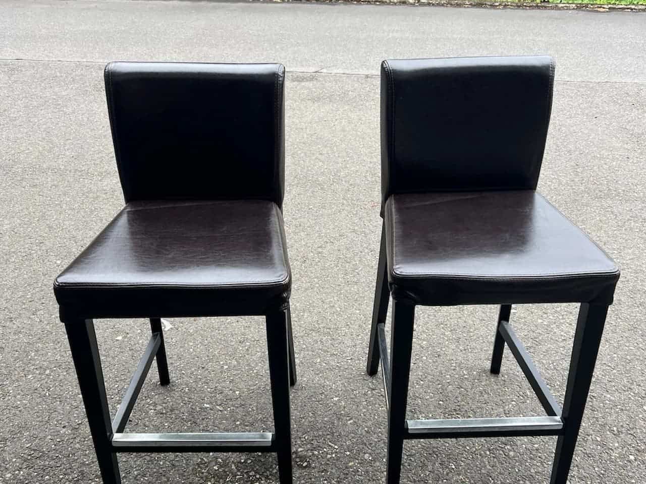 Two black leather bar stools with metal footrests and straight legs placed on an asphalt surface