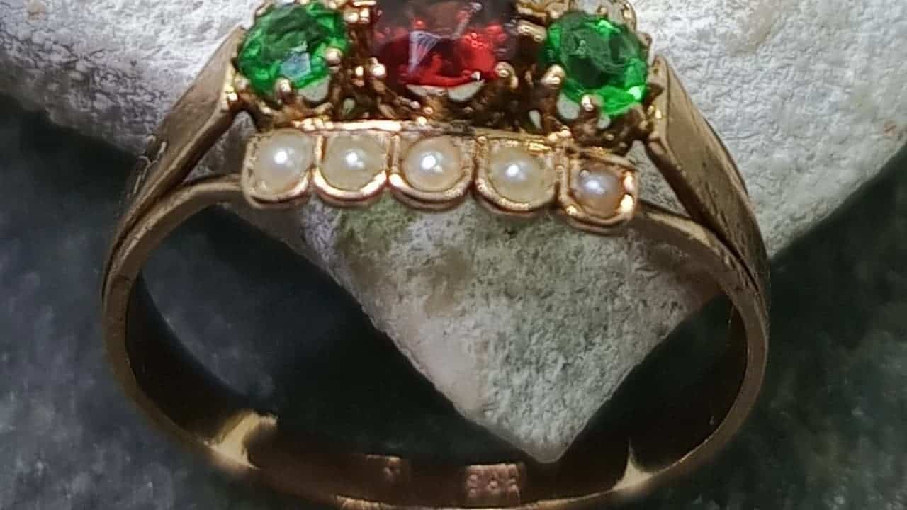 Victorian-era gold ring featuring emerald and ruby gemstones with row of pearls, displayed on textured stone surface