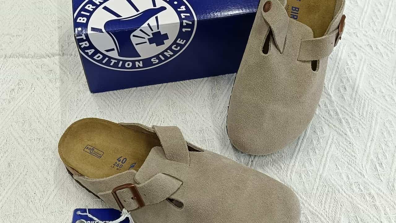 A pair of beige suede Birkenstock clogs with brown buckles, placed on a textured white surface beside a blue Birkenstock box