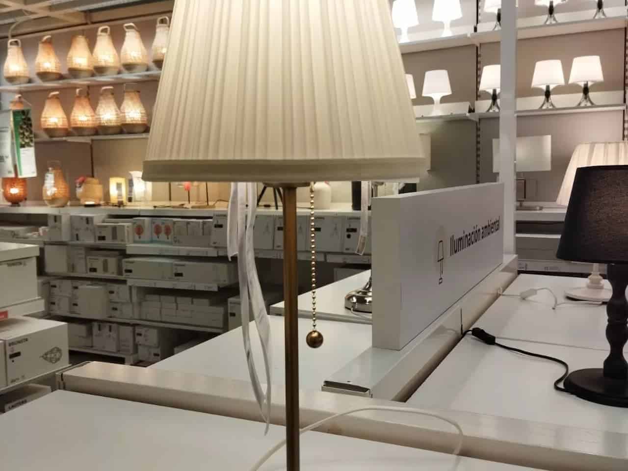 Beige table lamp with pleated fabric lampshade and brass pull-chain switch, displayed on a store counter among other lighting fixtures