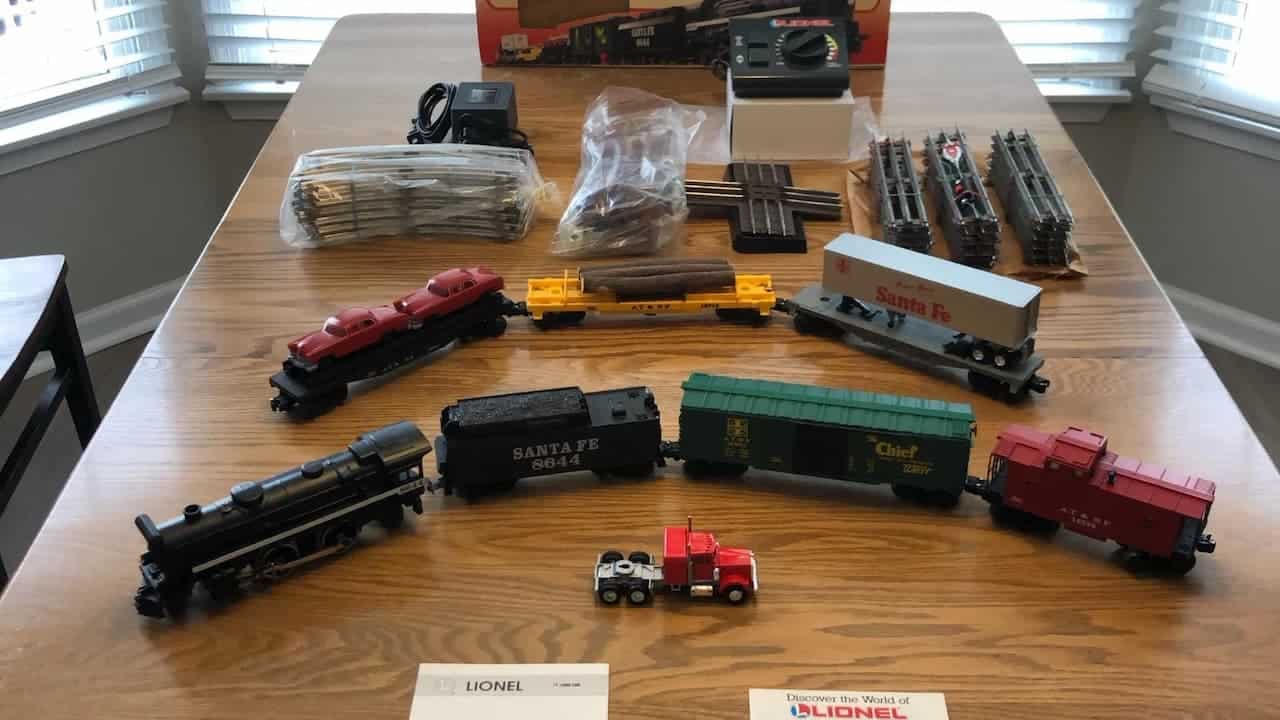 Lionel model train set arranged on a wooden table, including a black steam engine, Santa Fe coal car, boxcar, caboose, various freight cars, a red toy truck, power supply, track pieces, and controller