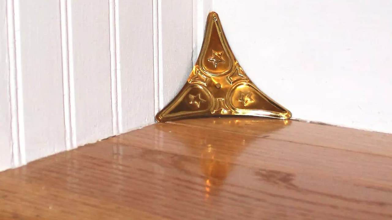 Golden triangular dust corner guard mounted where wooden stair step meets white wall baseboard in home interior