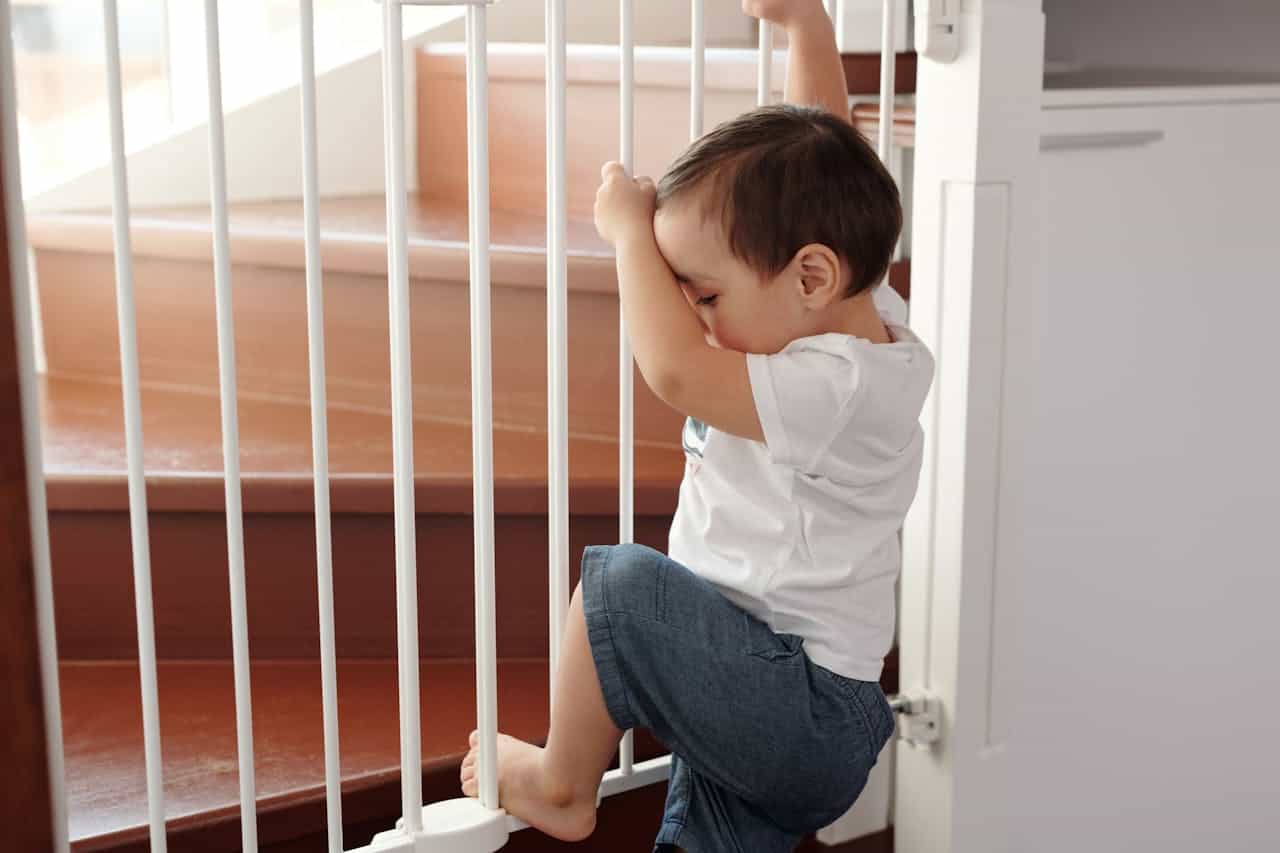 Child in white shirt and jeans sitting by white wooden safety gate, looking through the balusters