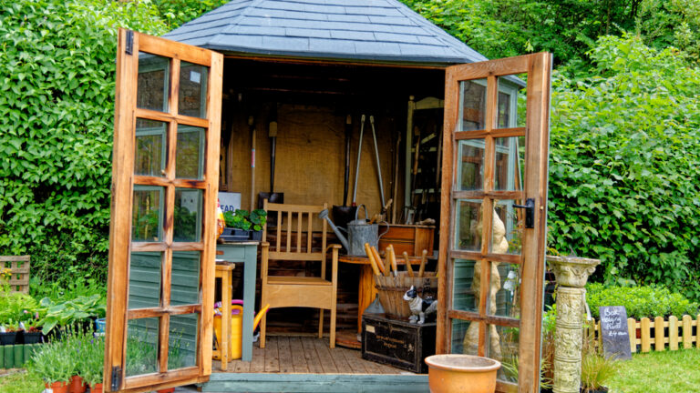 Garden shed with double glass-paneled doors wide open, revealing tools, benches, pots, and gardening supplies inside