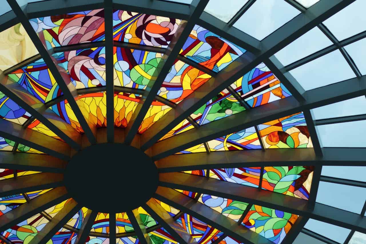A circular stained glass window with colorful geometric patterns radiating outward from a dark center, featuring vibrant blues, reds, yellows, and greens in triangular segments