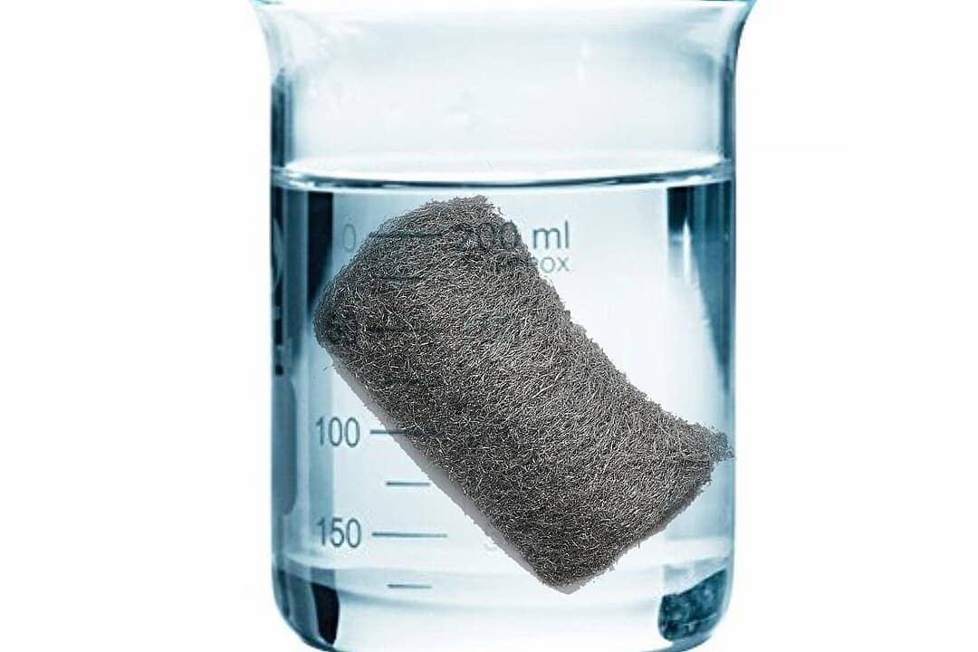 Gray steel wool pad floating in clear water inside glass laboratory beaker with measurement markings