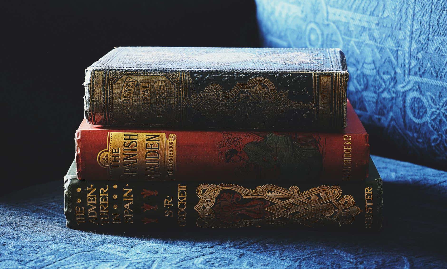 A_stack_of_antique_books_with_decorative_bindings_on_a_blue_fabric_sofa..Suzy_Hazelwood