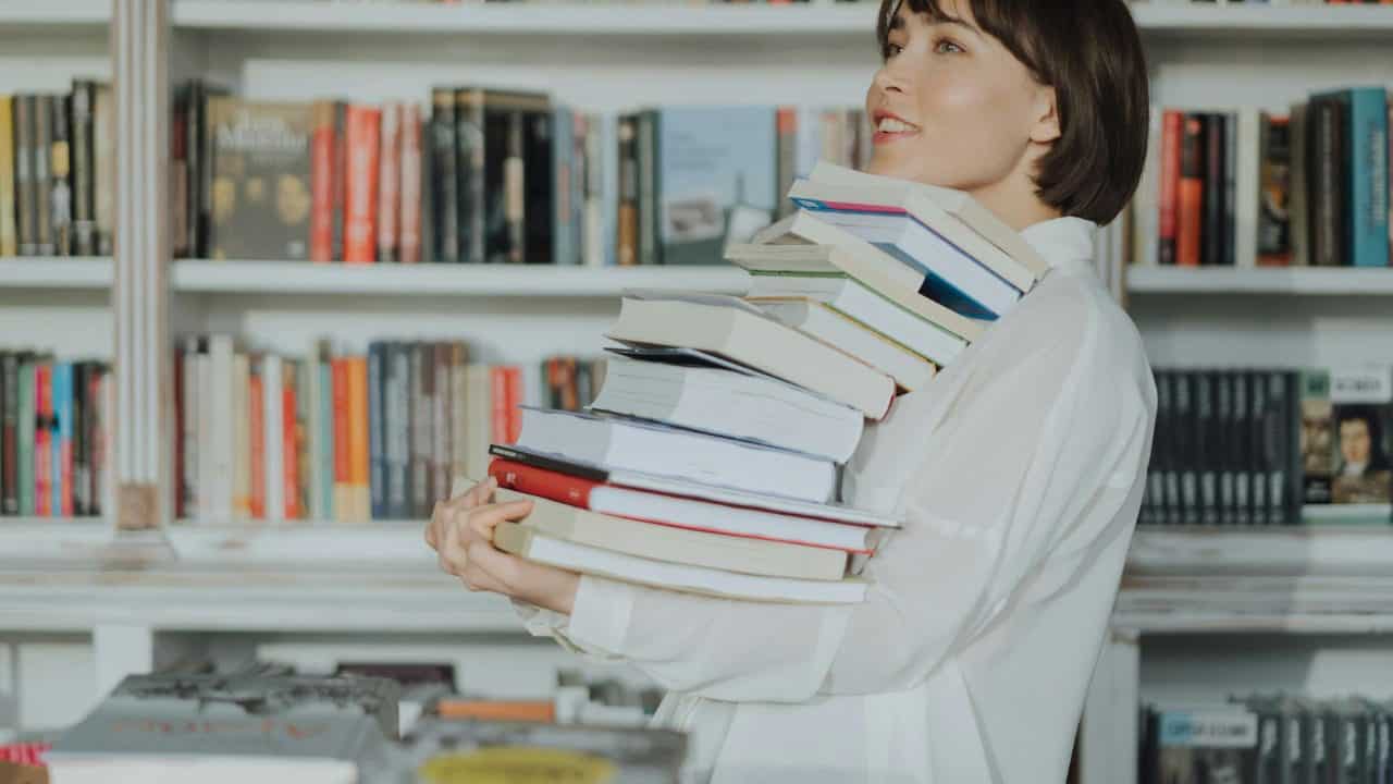 A woman joyfully holds a pile of books in a library, embodying the love of reading and learning.