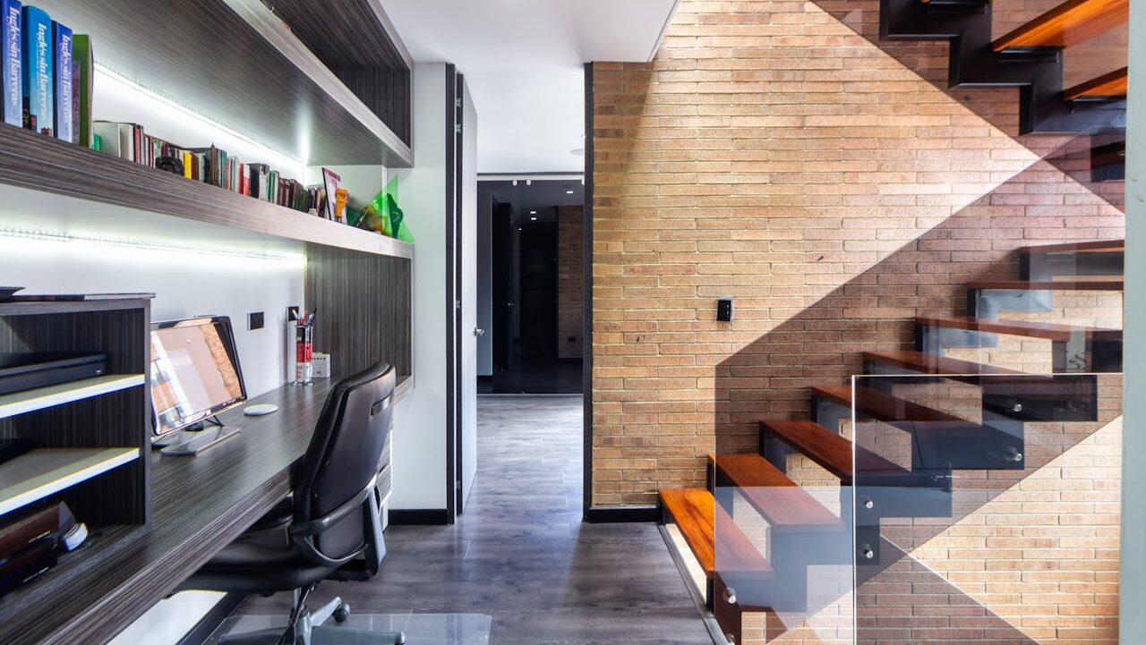 Modern workspace with desk, computer, black chair, bookshelves, wooden stairs with glass railing, and exposed brick accent wall