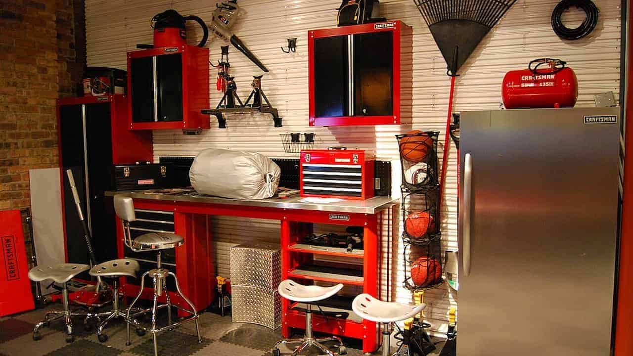 A well-organized garage workspace featuring red and black built-in Craftsman cabinets, a stainless steel workbench with tools and a toolbox, several industrial stools, a vertical sports ball rack, and a silver refrigerator