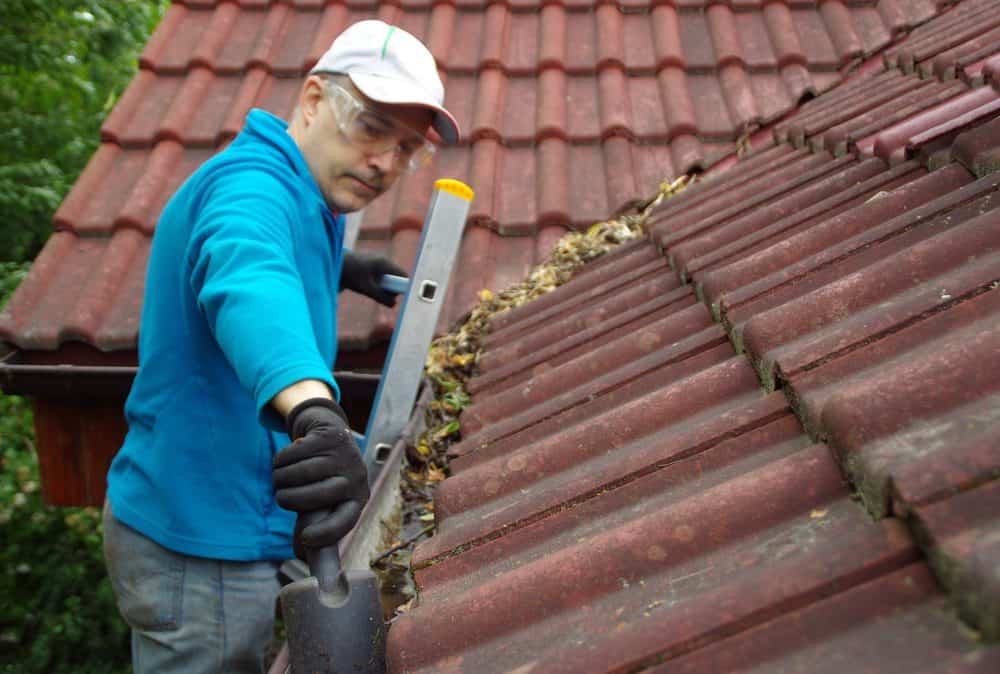 Man on ladder cleaning roof gutter filled with leaves on a red tiled rooftop