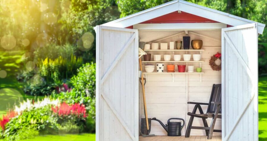 Garden shed filled with gardening tools. Shovels, rake, pots, water pitcher in storage hut. Green sunny garden in the background.