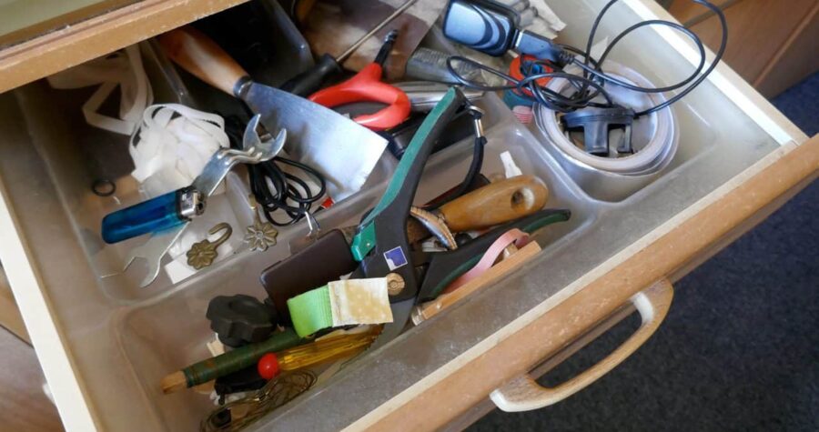 Cluttered wooden toolbox or drawer containing various tools including pliers, screwdrivers, and other hardware items haphazardly stored together. A blue checkmark appears in the corner