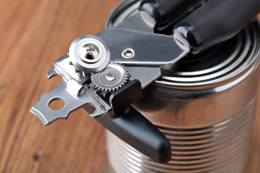 Manual can opener in use, cutting into a metal can, black handle, stainless steel cutting mechanism, placed on a wooden surface