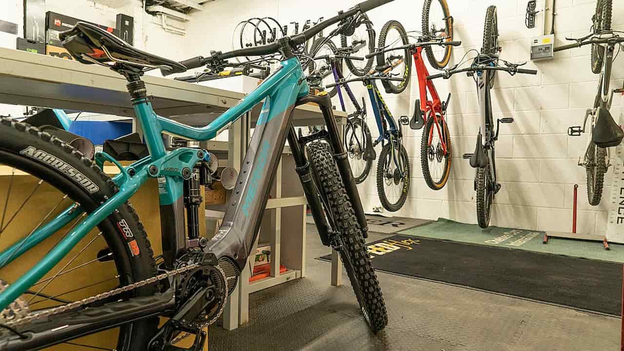 A bicycle workshop with several mountain bikes, including a teal and black full-suspension bike in the foreground and multiple bikes mounted vertically on the wall in the background