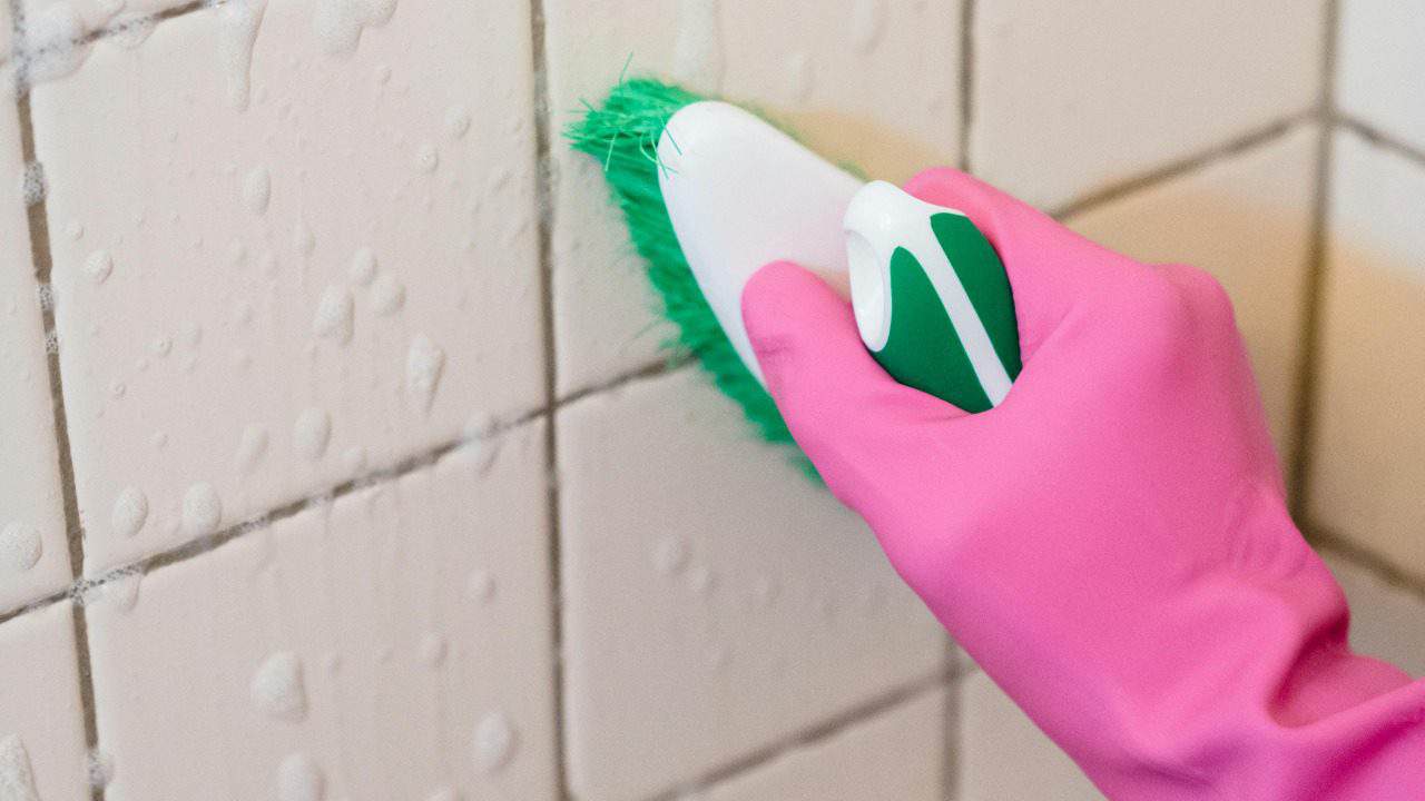 A hand in pink gloves scrubs bathroom tiles with a green brush to remove grime.
