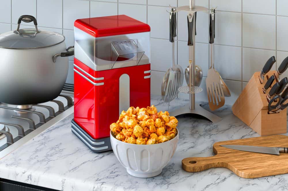 Red popcorn maker on a marble kitchen counter, freshly popped popcorn in a white bowl, stainless steel cookware in the background, wooden knife block with knives, kitchen utensils on a stand, tiled backsplash, cozy kitchen setup