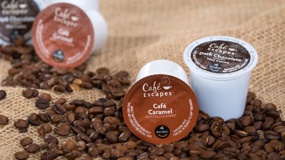 Café Escapes coffee pods, labeled Café Caramel and Dark Chocolate, surrounded by roasted coffee beans on burlap
