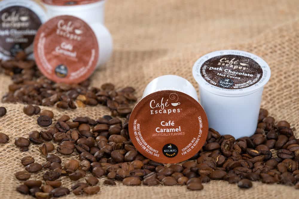 Café Escapes coffee pods, labeled Café Caramel and Dark Chocolate, surrounded by roasted coffee beans on burlap