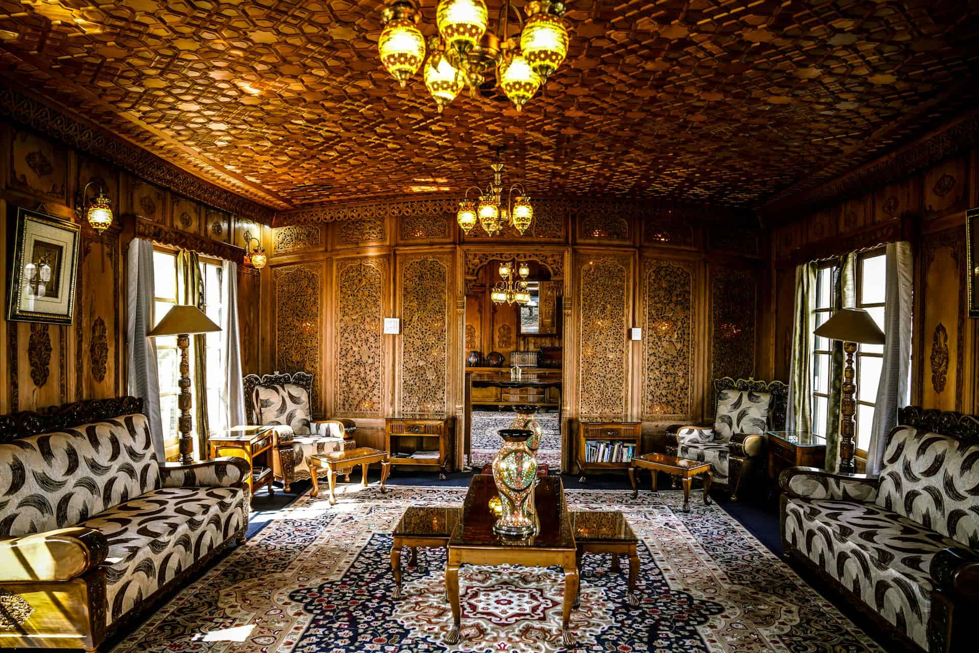 A traditionally furnished room with ornately carved wooden walls and ceiling, antique chandeliers, patterned rugs, classic armchairs, and a central wooden coffee table with a decorative vase