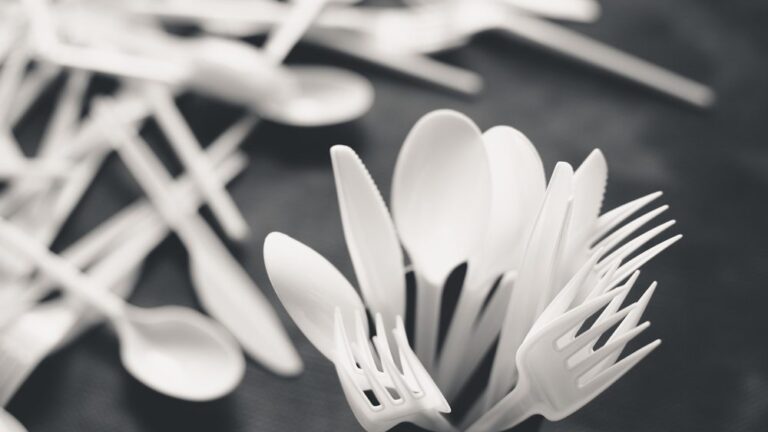White plastic forks, spoons, and knives arranged in a container, with more scattered on a dark surface in the background