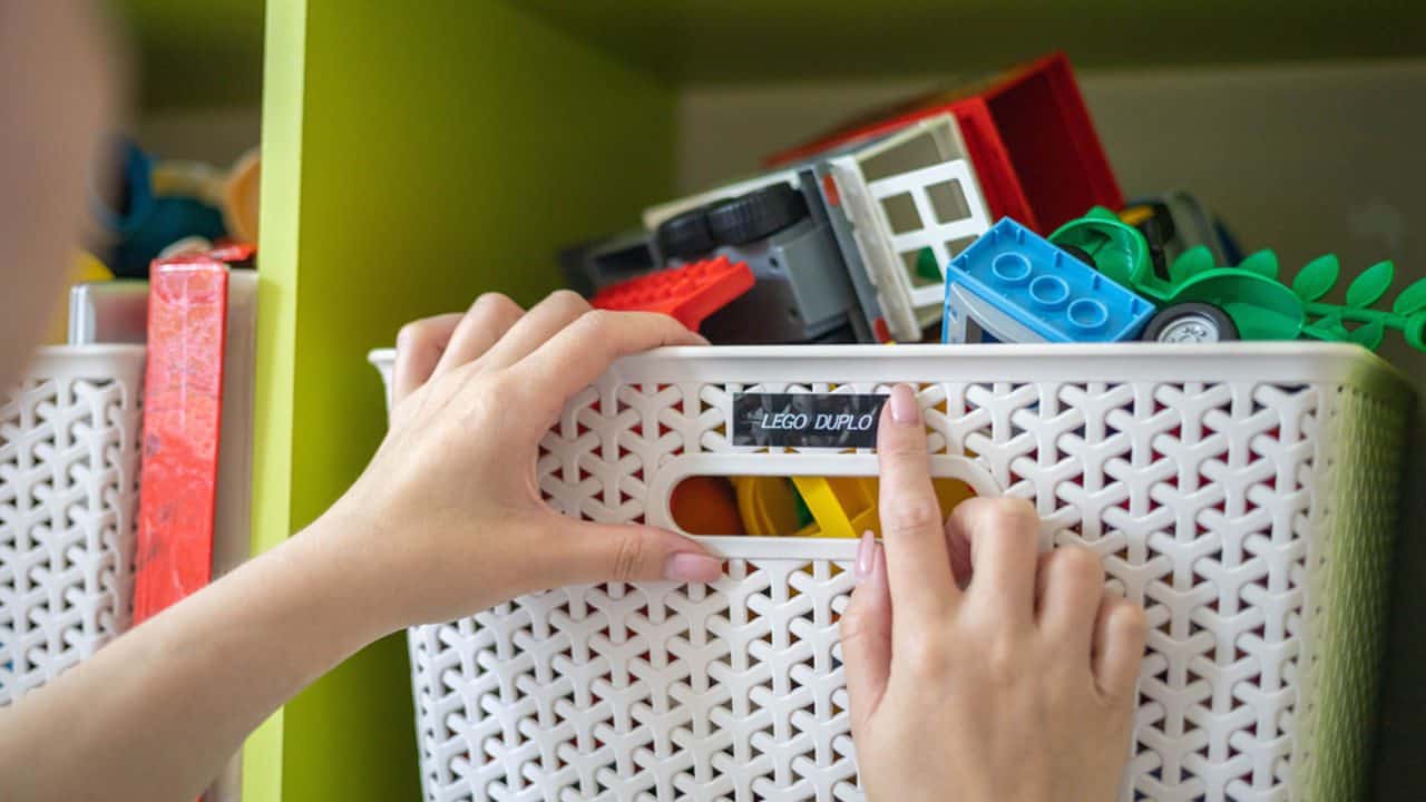 Hands placing or pulling a white perforated storage bin labeled “cars and trucks,” filled with toy vehicles and building blocks, against a green wall or shelf background