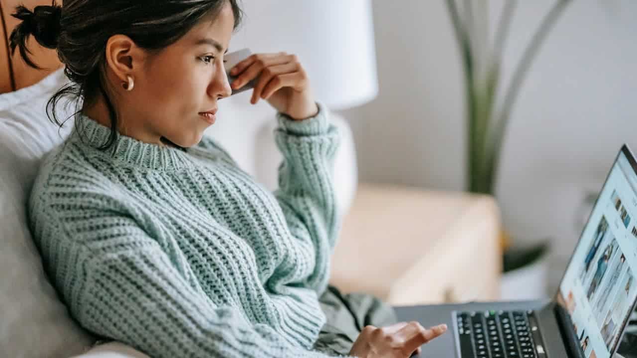 woman in mint green sweater, sitting on bed, using laptop, focused expression, online shopping website on screen, indoor setting with lamp and plant in background