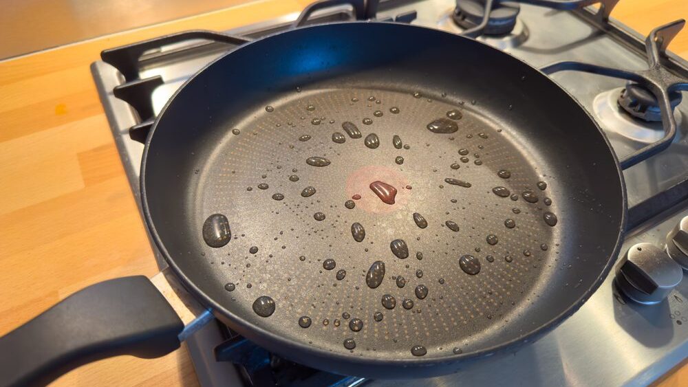 breaded food item into a frying pan, stovetop cooking, nonstick pan in use, wearing a green apron, kitchen background blurred.