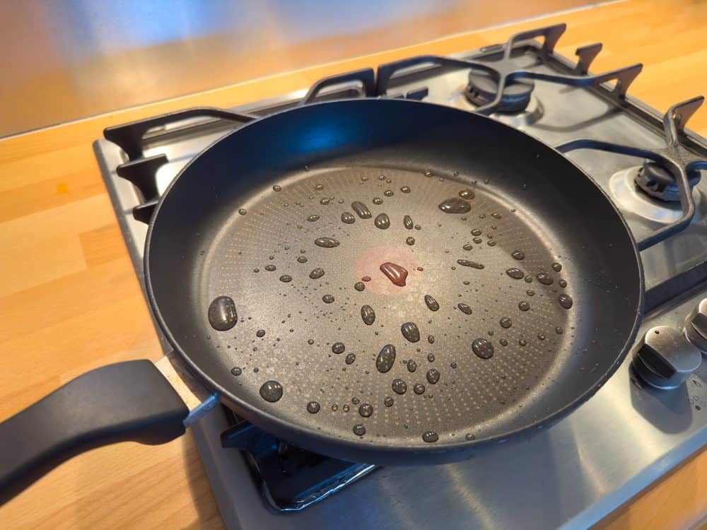 breaded food item into a frying pan, stovetop cooking, nonstick pan in use, wearing a green apron, kitchen background blurred.