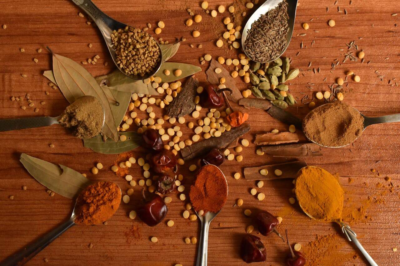 Assorted ground and whole spices on wooden surface, spoons holding turmeric, chili powder, cumin, cinnamon, coriander, cardamom, and fenugreek