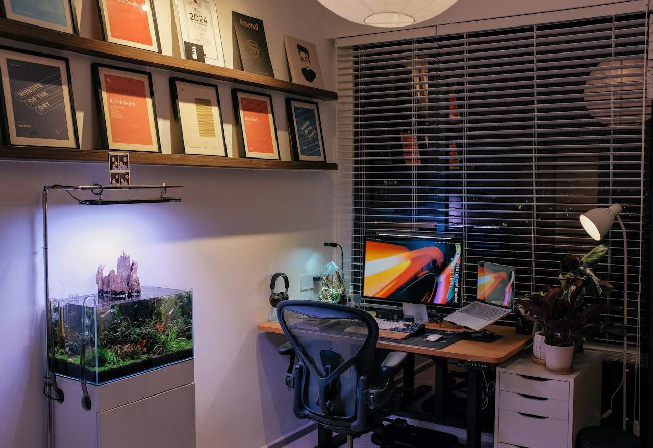 Modern home office setup, dual monitors on wooden desk, ergonomic chair, fish tank with plants, wall-mounted awards and certificates, window blinds, and ambient lighting