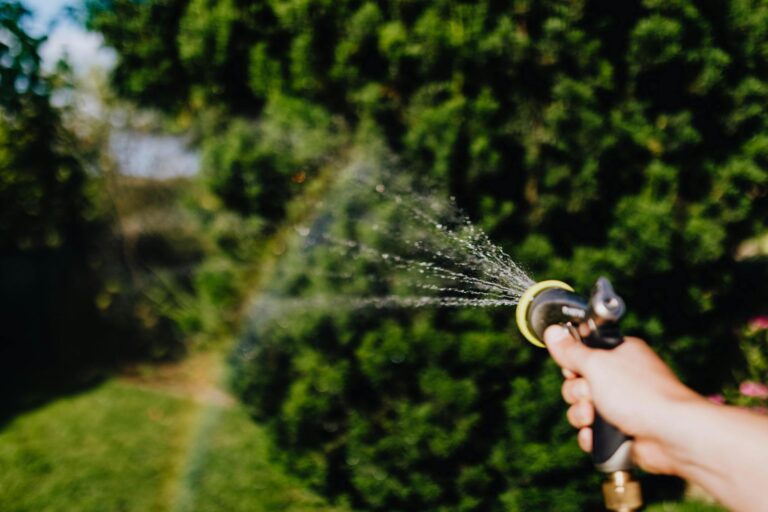 A hand holding a hose watering plants in a lush garden setting.